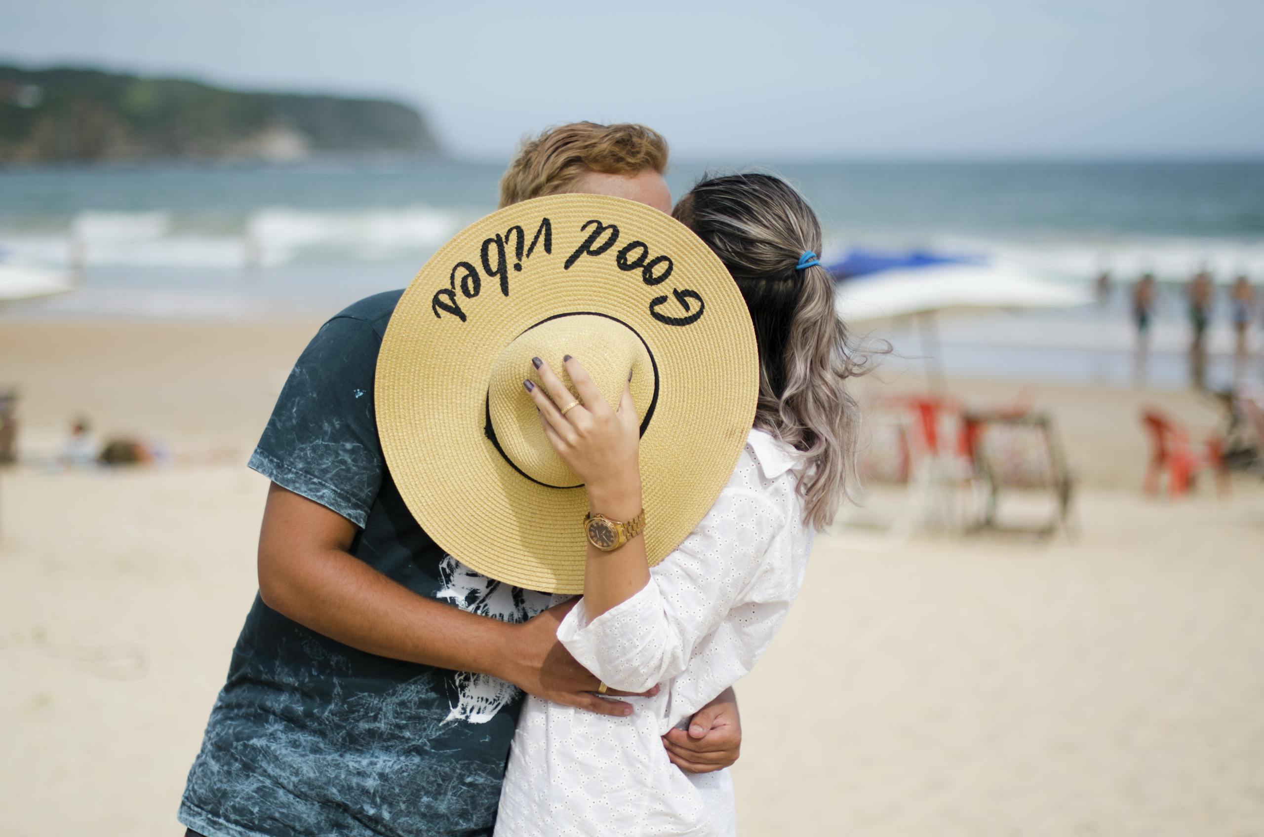 A couple shares a romantic kiss on a sunny beach, hidden behind a wide-brimmed hat.